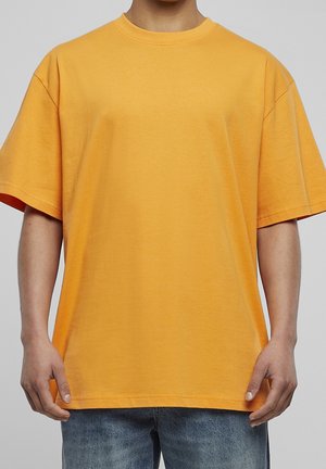 Person wearing a plain oversized short-sleeve orange t-shirt and blue denim jeans, standing against a neutral gray background.