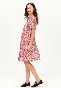 Rode bloemenjurk met korte mouwen en een uitlopende rok. Bevat blauwe en groene bloemmotieven. G gedragen met zwarte loafers.