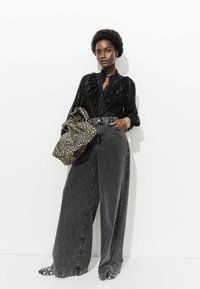 Blouse noire à volants, jean gris taille haute à jambes larges, et sac à imprimé léopard. Les chaussures présentent un motif floral noir et blanc.