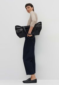 Borsa a tracolla nera con pannelli in rete e manici piatti, con un design minimale. Indossata con una maglietta beige e pantaloni in denim scuro.
