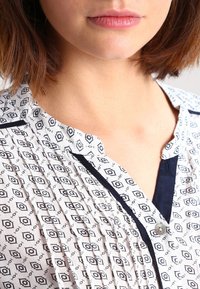 Blusa bianca con un motivo geometrico nero, bordino blu navy lungo il collo e la patta dei bottoni, con una texture plissettata.