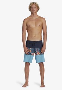 Shorts de bain avec une taille bleu foncé, motif floral en orange et vert, et une partie inférieure bleu clair. Fabriqués en tissu à séchage rapide.