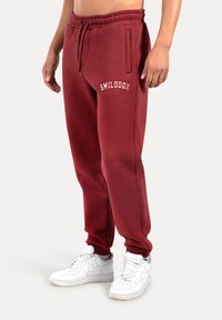 Bordeaux kleurige sweatpants met een elastische tailleband en trekkoord, voorzien van een witte "SMILODOX" logo op de bovenste linker dij. Gladde textuur.