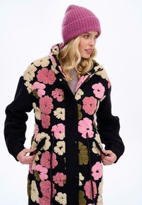 Fleecový kabát s černým základem a barevnými květinovými vzory v růžové, krémové a zelené barvě. Má zapínání na druky a boční kapsy.