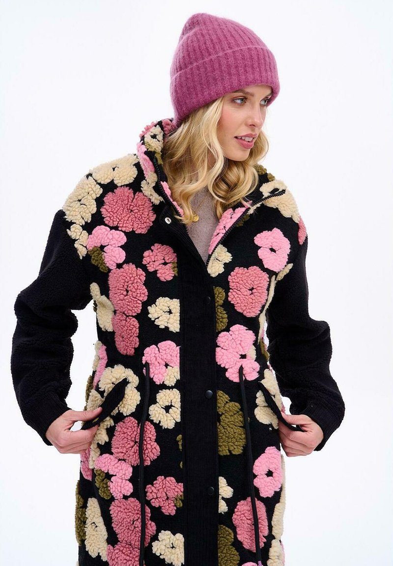 Fleecový kabát s černým základem a barevnými květinovými vzory v růžové, krémové a zelené barvě. Má zapínání na druky a boční kapsy.