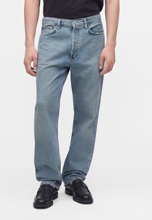 Mand iført lyseblå jeans med lige ben og sorte læder loafers mod en ensfarvet hvid baggrund.