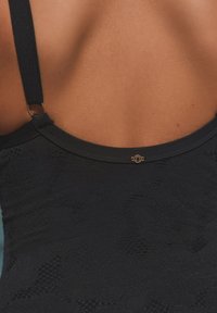 Must ujumisriiet tekstuurse mustriga, reguleeritavate rihmadega ja kuldse detailiga seljal, rõhutades sujuva kangasegu viimistlust.