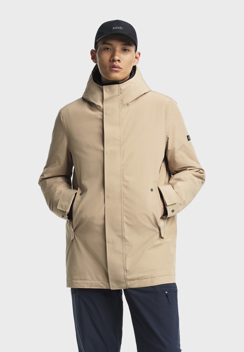 Beige geïsoleerde jas met een hoge kraag, voorkant met drukknoopsluiting en zijzakken. Gestyled met een zwarte pet, gedragen met donkere broek.