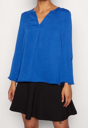 Kobieta ubrana w niebieską bluzkę z długim rękawem i dekoltem w serek oraz czarną, rozkloszowaną spódnicę do połowy uda na gładkim jasnym tle.