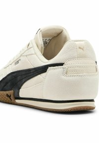 Zapatilla de ante blanca con franja lateral de cuero negro, talón texturizado y suela de goma. Presenta un branding sutil y elementos de diseño minimalistas.