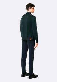 Felpa verde scuro con colletto alto, spacchi laterali e un accento arancione. Abbinata a pantaloni scuri e scarpe nere, vista da dietro.