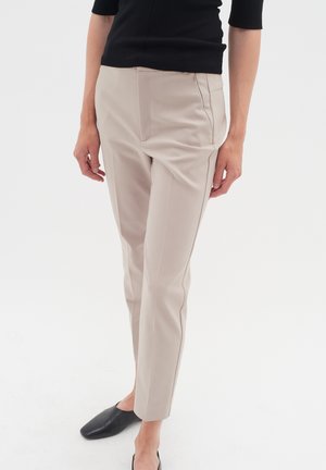 Pantalon beige slim-fit raccourci porté par une personne en haut noir et chaussures plates noires, debout devant un fond blanc uni.