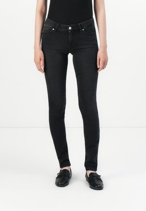 Slim-fit black jeans worn with black slip-on loafers and a tucked-in black top, shown against a plain white background.