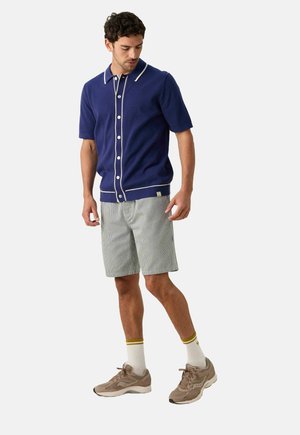 Man in een blauwe kortarmige blouse, gestreepte shorts, witte sokken met gele strepen en beige hardloopschoenen, staat en kijkt naar beneden.