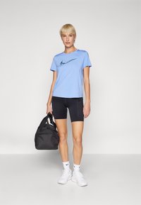 Camiseta deportiva de color azul claro con un logo negro de Nike, combinada con pantalones cortos negros y zapatillas blancas. Se sostiene una bolsa de deporte negra con una mano.