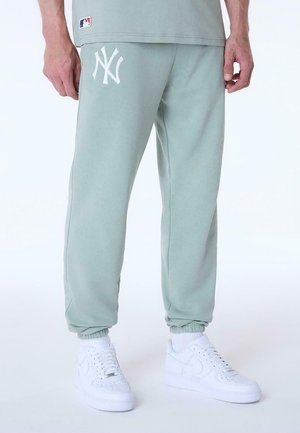 Persona que lleva pantalones deportivos verde claro con el logo blanco de los New York Yankees y zapatillas blancas, de pie con las manos relajadas.