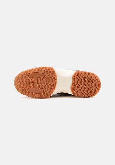 Semelle de chaussure avec grip en caoutchouc marron, motif ondulé et une grande forme "H" dans la partie avant de la semelle sur un fond de semelle intermédiaire blanche.