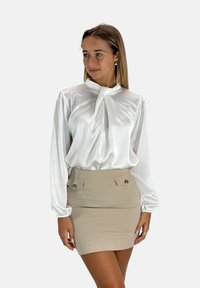 Witte satijnen blouse met lange mouwen en een strik bij de hals, gecombineerd met een beige aansluitende mini-jurk met twee voorkeurszakken en gouden accenten.