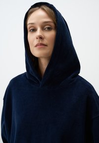 Marineblauwe hoodie gemaakt van een textuurlijke stof, met een losse pasvorm en een grote capuchon, zonder zichtbare logo's of versieringen.