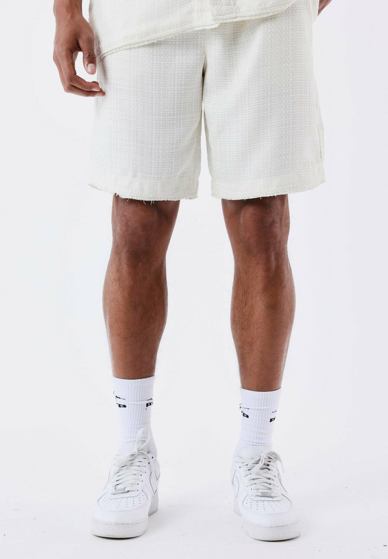 Shorts blancos texturizados con dobladillos deshilachados, combinados con calcetines blancos y zapatillas. Los shorts presentan un patrón tejido y un ajuste relajado.