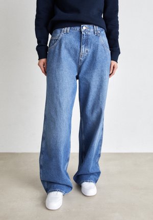 Persona con vaqueros azules holgados, zapatillas blancas y una camiseta oscura de manga larga, de pie sobre un suelo neutro.