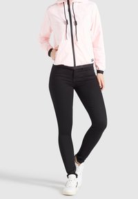 Jasnoróżowa kurtka zapinana na zamek z kapturem i czarnymi akcentami, w zestawieniu z czarnymi, dopasowanymi dżinsami i białymi butami sportowymi. Gładka tekstura.