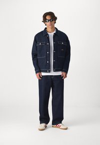 Jaqueta de ganga escura com costuras claras, bolsos frontais e botões de metal. Combinada com jeans escuros soltos e sapatos de creme com riscas vermelhas.