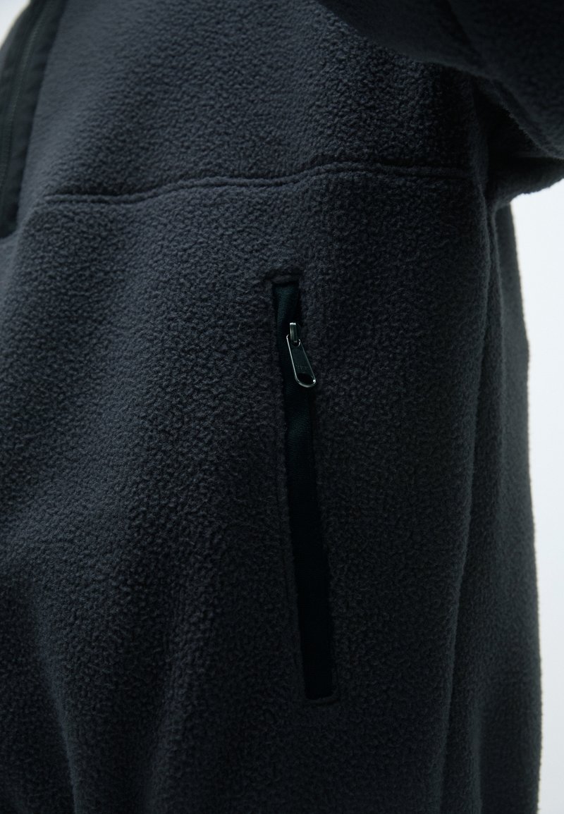 Svart fleecjacka med en texturerad yta, en ficka med dragkedja och ett smidigt tygfoder. Intrikata sömmar längs sömmarna.