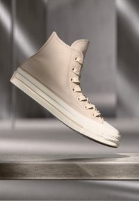 Høye sneakers i lys beige lerret med hvit gummisåle, med øyer, snører og en teksturert tåhette. Flyter mot en nøytral bakgrunn.