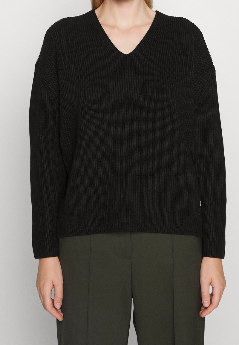 Czarny dziany sweter z dekoltem w serek, długimi rękawami i prążkowaną fakturą. Luźny krój, zestawiony z oliwkowozielonymi spodniami.