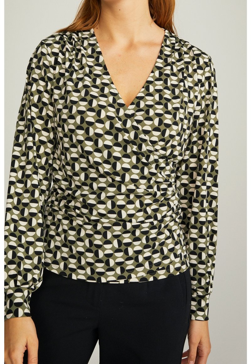 Langärmliges, wickelfront Top in Grün mit geometrischen Mustern in Schwarz, Weiß und Creme. Weicher Stoff mit plissierten Details um die Taille.