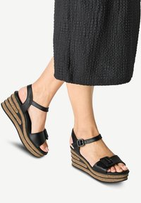 Černé kožené klínové sandály mají široký pásek s přezkou, strukturovanou plošinu podobnou korku a otevřenou špičku pro lepší prodyšnost.