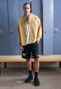 Yellow denim shirt, white T-shirt beneath, black shorts with yellow print, black clogs, and a patterned scarf tied around the neck.