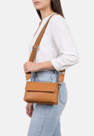 Donna che indossa una camicetta bianca e jeans azzurri chiari porta una borsa a tracolla in pelle marrone con tracolla larga regolabile.
