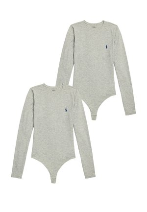 Femme debout portant un body gris à manches longues avec un pull gris noué autour de la taille et des chaussettes en tricot crème sur fond blanc.