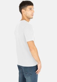 Weißes Kurzarm-T-Shirt mit Rundhalsausschnitt, seitlich betrachtet, kombiniert mit blauen Jeans, die zwei Gesäßtaschen und sichtbare Nähte aufweisen.