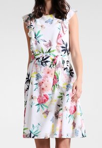 Robe fleurie avec des manches volantées, fond blanc, pivoine rose et verte, papillons et feuilles ; ceinturée à la taille ; longueur genoux.