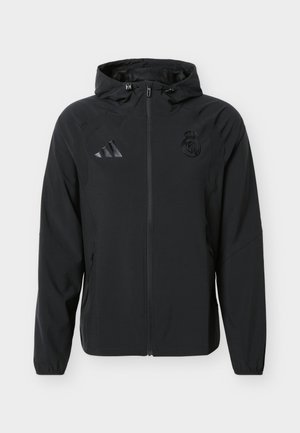 Czarna kurtka z kapturem zapinana na pełny zamek, z logo Adidas na prawej piersi oraz herbem Realu Madryt na lewej piersi.