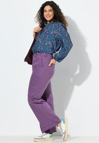 Jeans de pierna ancha morados combinados con una blusa de color azul oscuro con estampado. La modelo lleva zapatillas blancas y en tonos pastel, sosteniendo un bolso oscuro.