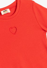Rotes, geripptes Kurzarm-T-Shirt mit Rundhals-Ausschnitt, das ein gesticktes Herzdetail auf der Vorderseite aufweist. Hergestellt aus weichem, strukturiertem Stoff.