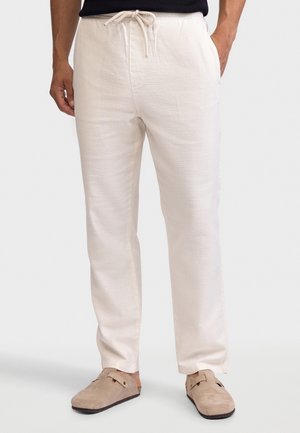 Muž v nohaviciach sťahovaných šnúrkou v bielej farbe s bočnými vreckami a béžových slip-on topánkach, stojaci s rukami v bodoch.