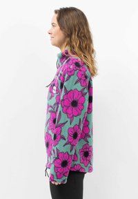 Fleecejacke mit türkisfarbener Basis und großem pinkfarbenem Blumenmuster, ausgestattet mit einem hohen Kragen und einem Halbzippverschluss, verstellbarem Saum und seitlichen Taschen.