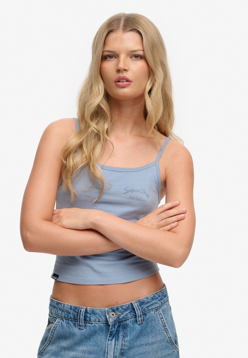 Mujer rubia con cabello ondulado lleva una camiseta sin mangas azul claro y pantalones vaqueros, de pie con los brazos cruzados frente a un fondo blanco.