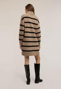 Béžové pletené šaty s černými vodorovnými pruhy, které mají vysoký límec a dlouhé rukávy, doplněné černými lesklými kozačkami ke kolenům.