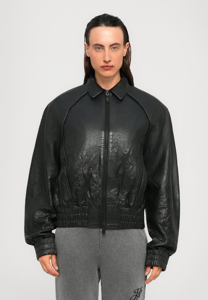 Persona con capelli pettinati all'indietro, che indossa una giacca nera in pelle con zip e pantaloni grigi con iniziali ricamate.