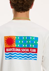 Fehér, hosszú ujjú ing, mely multicolor grafikát tartalmaz, piros nappal, hullámmintával és vastag betűkkel írt "BARCELONA SOCIAL CLUB" felirattal.