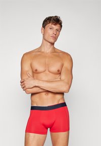 Boxer-briefs rouges avec une ceinture élastique bleu marine arborant un logo, fabriqués à partir d'un tissu lisse avec un design ajusté.