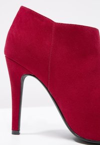 Bottine à talon haut en daim rouge avec un bout pointu et un talon stilettos noir lisse. Détails de coutures le long du côté.