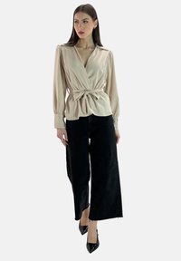 Blusa avvolgente beige con un nastro in vita, maniche lunghe e colletto, abbinata a jeans neri a Vita alta e larghi e tacchi neri.