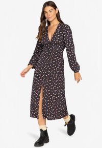 Femeie îmbrăcată într-o rochie midi florală neagră cu mâneci lungi, fendue în față, șosete crem și bocanci negri, stând pe un fundal alb.